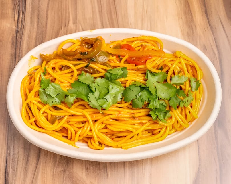 A white oval bowl filled with yellow spaghetti, bell peppers, and fresh cilantro on a wooden table.