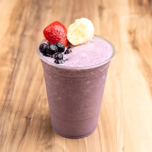 Purple berry smoothie in a clear cup, topped with blueberries, a strawberry, and banana slices.