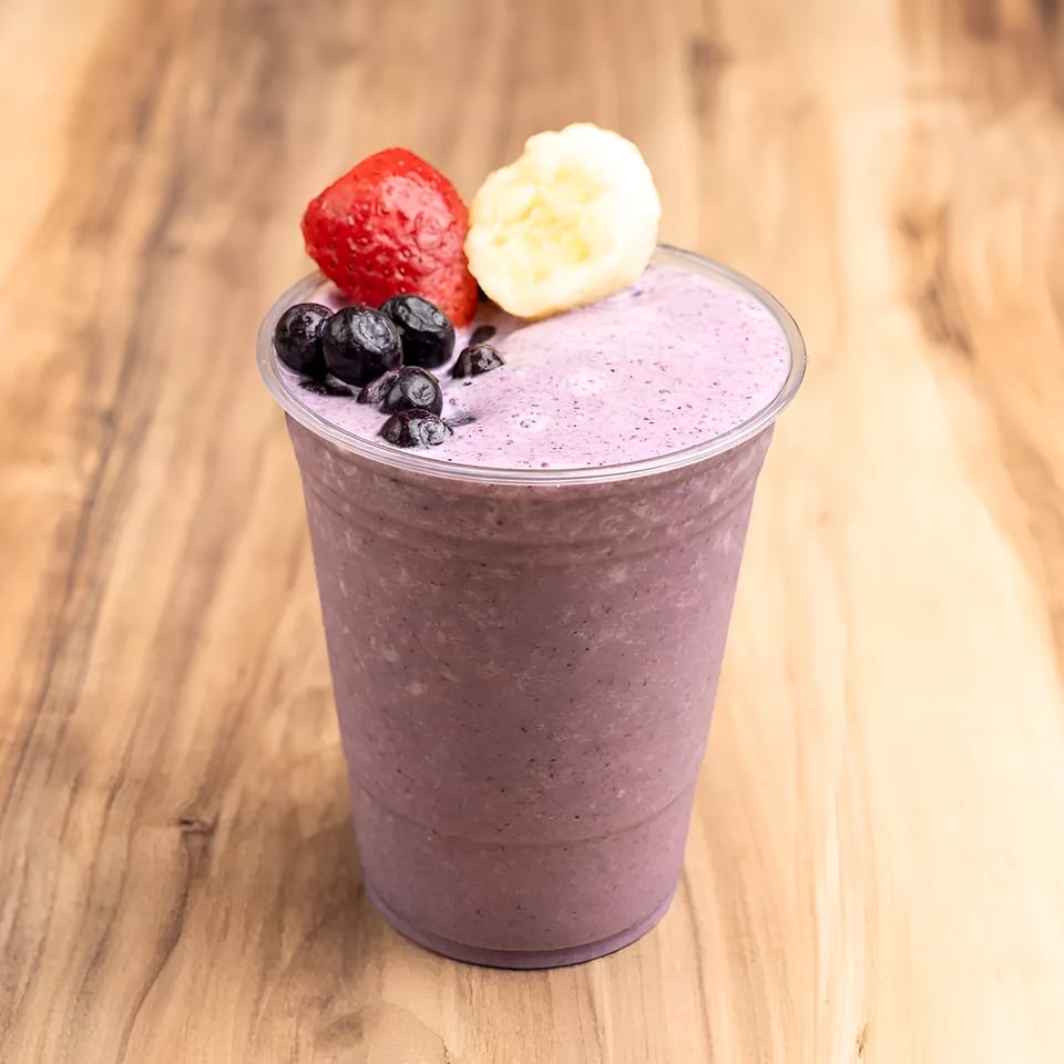 Purple berry smoothie in a clear cup, topped with blueberries, a strawberry, and banana slices.