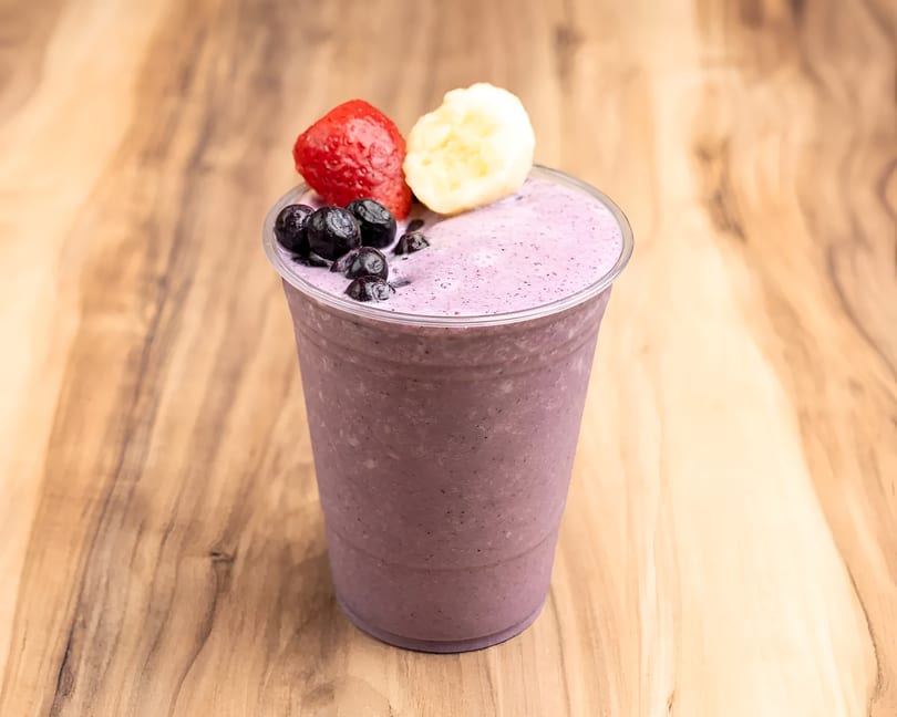Purple berry smoothie in a clear cup, topped with blueberries, a strawberry, and banana slices.