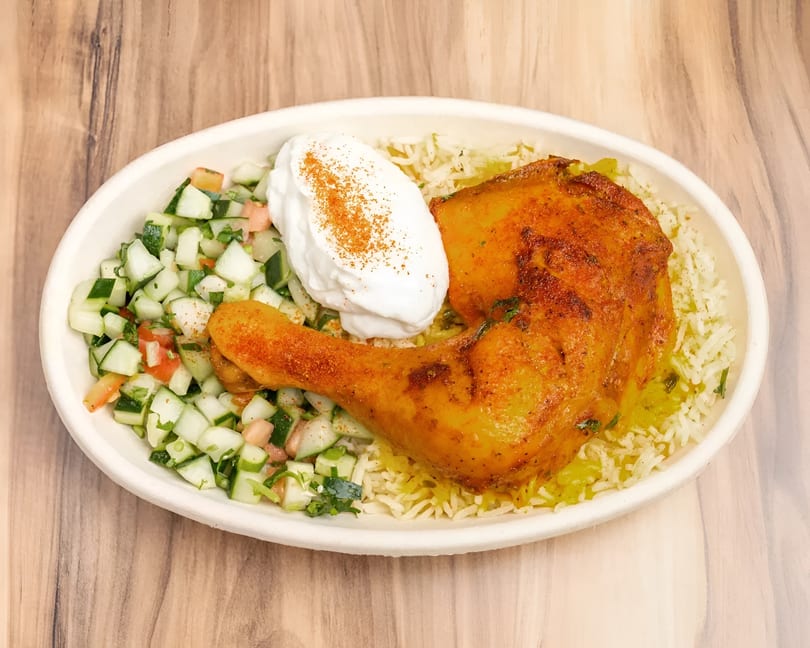 A chicken leg with rice, cucumber salad, and a dollop of sour cream in a white bowl.