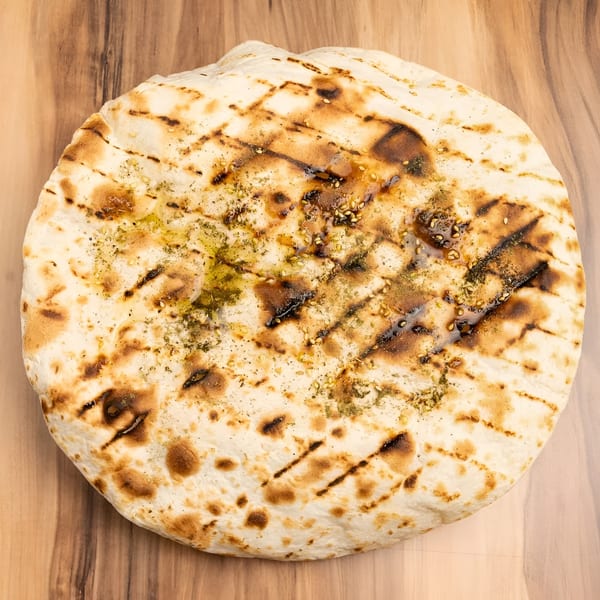 Grilled flatbread with olive oil, herbs, and sesame seeds on a wooden surface.