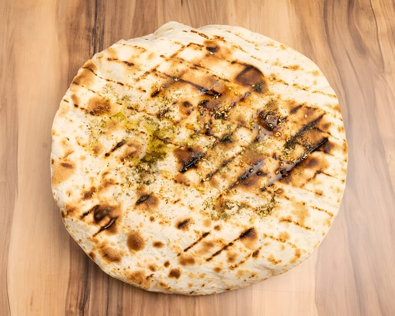 Grilled flatbread with olive oil, herbs, and sesame seeds on a wooden surface.
