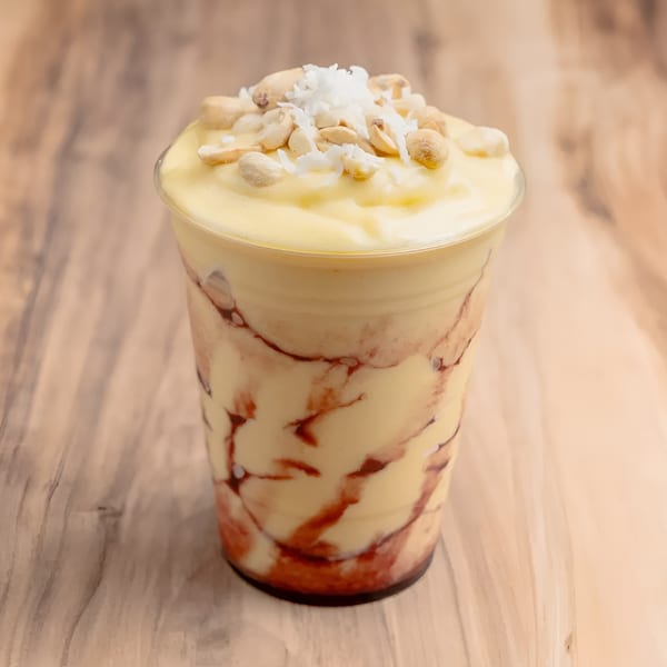 A plastic cup of yellow mango smoothie with red swirls, topped with peanuts and shredded coconut.