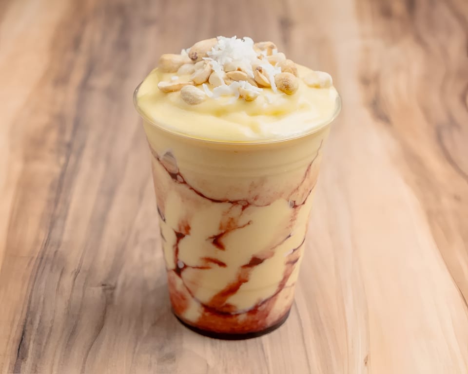 A plastic cup of yellow mango smoothie with red swirls, topped with peanuts and shredded coconut.