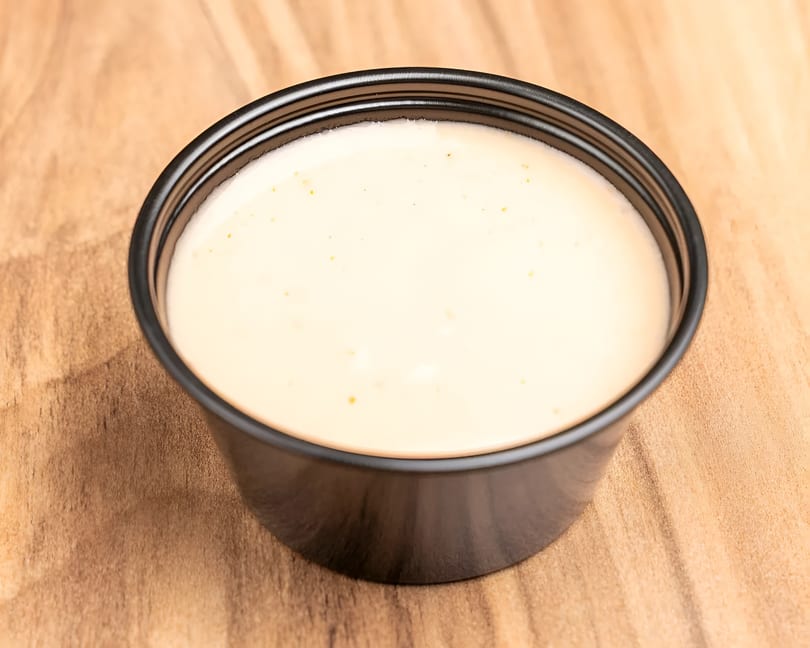 Creamy white sauce with small orange specks in a black plastic cup on a wooden surface.