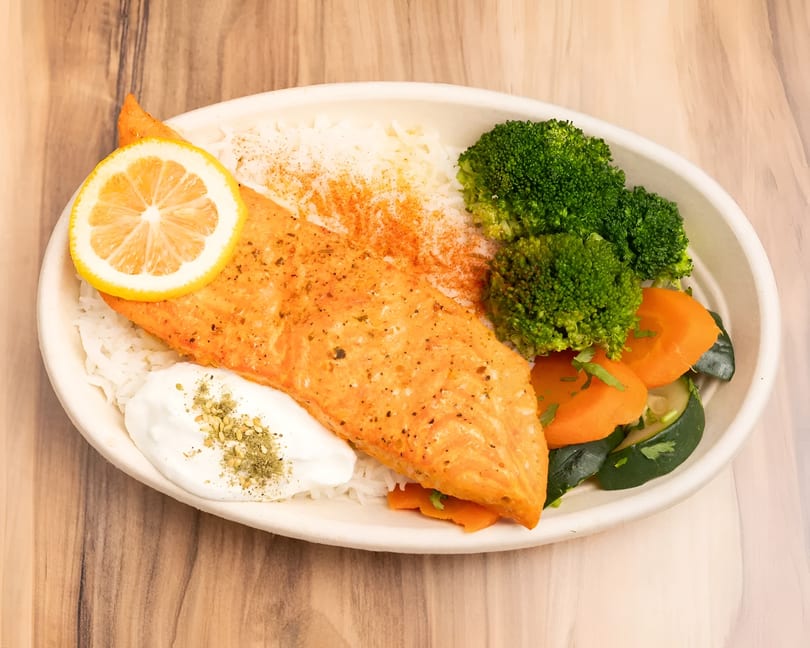 Salmon fillet with rice, broccoli, carrots, zucchini, a lemon slice, and a dollop of sauce.