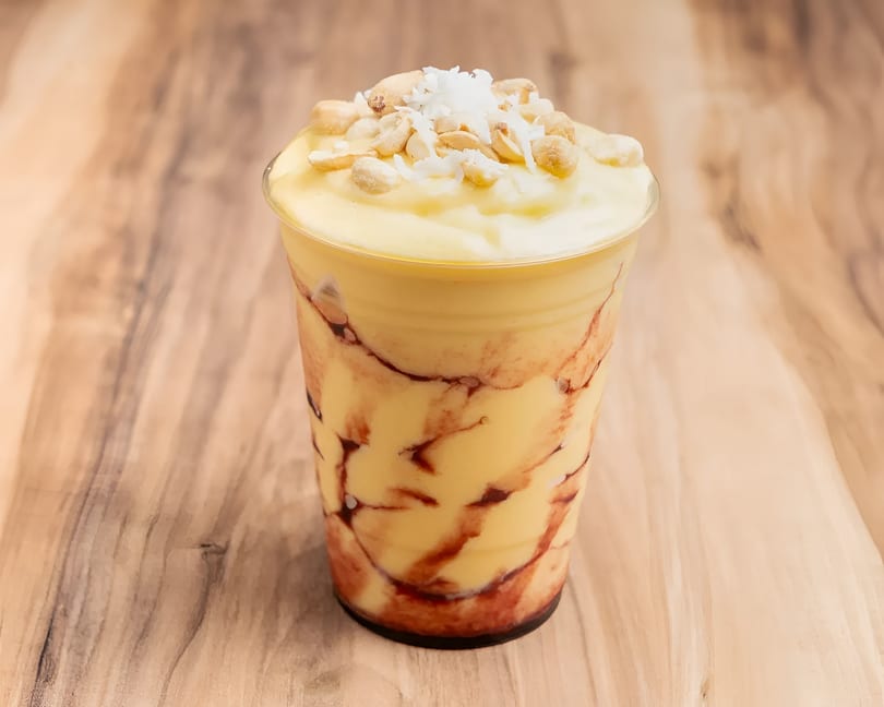 A yellow smoothie with red swirls, topped with peanuts and shredded coconut, on a wooden table.