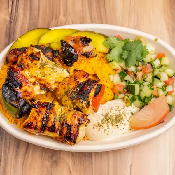 Grilled chicken and vegetable skewers with yellow rice, hummus, cucumber salad, and pickles.