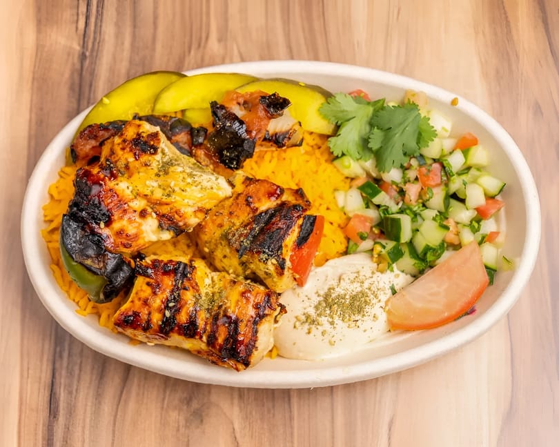 Grilled chicken and vegetable skewers with yellow rice, hummus, cucumber salad, and pickles.